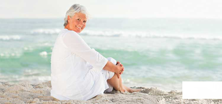 Se ve una mujer riendo con pelos blancos. Está sentada en la playa y disfruta de unas buenas vistas al mar.